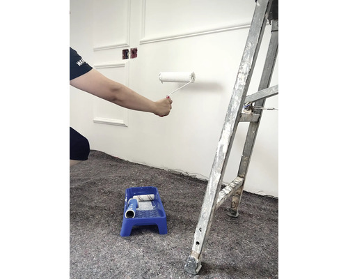 Person målar vägg med färgroller, färgtråg och stege i rummet.