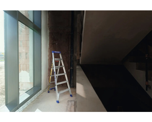 En stege står i ett oinrett hus bredvid ett fönster.