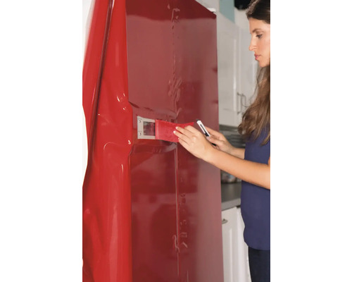 Kvinna applicerar en röd möbelfolie på ett kylskåp med en skrapa och en kniv.