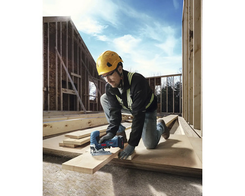 En hantverkare med skyddshjälm och skyddsglasögon sågar en träplanka med en sticksåg på en byggarbetsplats.