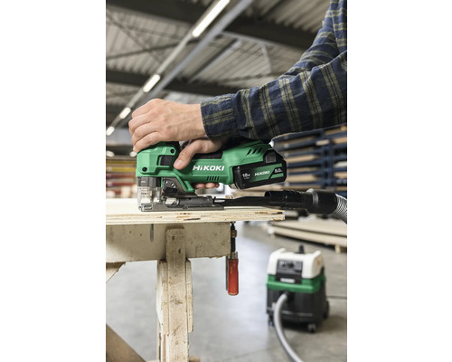 Person använder en grön Hikoki sladdlös sticksåg på en träyta med en dammsugare ansluten.