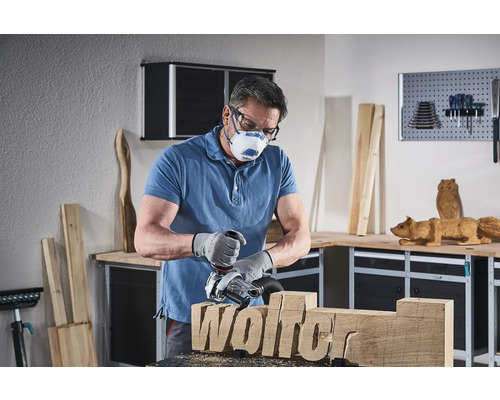 Man arbetar med en träfräs på en träbit med inskriptionen Wolfer, bär skyddsmask och skyddsglasögon i en verkstad.