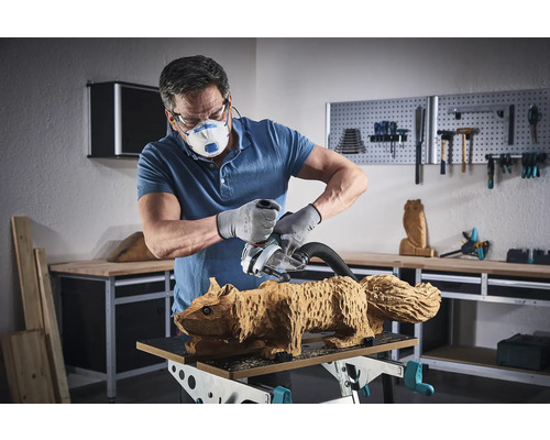 Man slipar en ekorrfigur i trä med en slipmaskin. Han bär mask, skyddsglasögon och handskar.