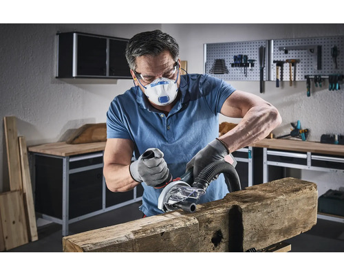 En man slipar en träbit med ett elverktyg i en verkstad, med ett skyddsmask och handskar på.