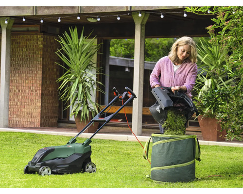 En kvinna tömmer gräs i en trädgårdssäck bredvid en gräsklippare på en gräsmatta.