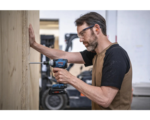 En man skruvar med en batteridriven mutterdragare en skruv i en träplanka. Han bär skyddsglasögon och öronproppar. Bosch logotyp.