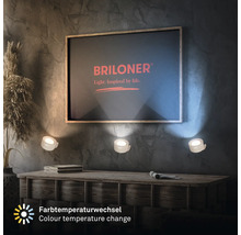 Vardagsrum med byrå, dekoration och tre vägglampor under en bild med Briloner-logotypen. Lamporna avger ett varmt ljus och erbjuder färgförändring.