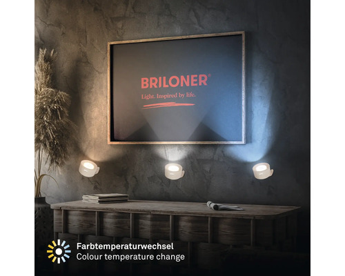 Vardagsrum med byrå, dekoration och tre vägglampor under en bild med Briloner-logotypen. Lamporna avger ett varmt ljus och erbjuder färgförändring.