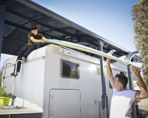 Två personer lyfter en surfbräda på en husbil med markis.