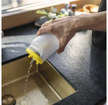 En person häller vatten från en vit plastbehållare med gult lock i en guldfärgad diskho.
