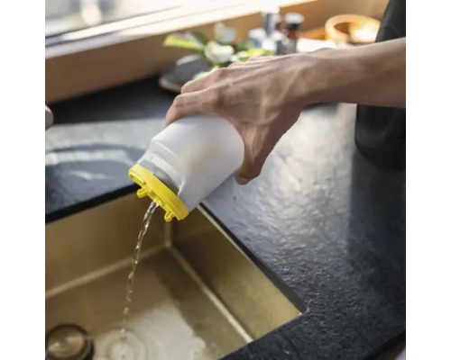 En person häller vatten från en vit plastbehållare med gult lock i en guldfärgad diskho.