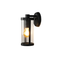 Modern utomhusvägglampa i cylindrisk form av svart metall och klart glas med tänd ljuskälla.