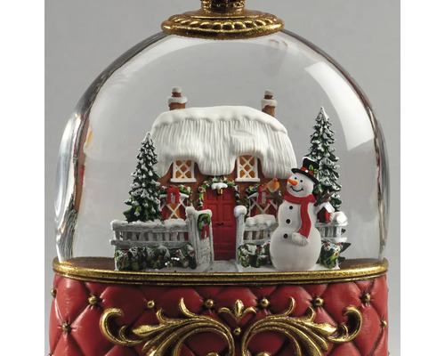 Juldekoration snökula med snötäckt hus, snögubbe och granar på en röd bas med guldfärgade detaljer.