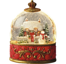 Snöglob med snötäckt hus, snögubbe och granar på en röd sockel med en krona överst.