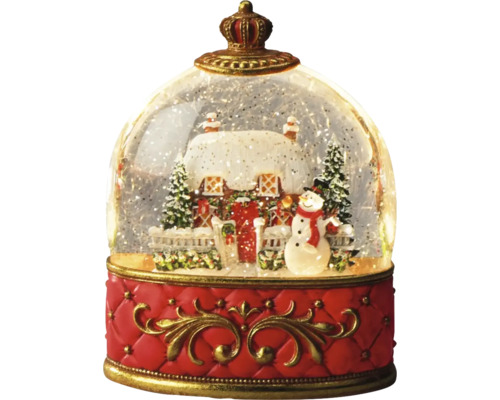 Snöglob med snötäckt hus, snögubbe och granar på en röd sockel med en krona överst.