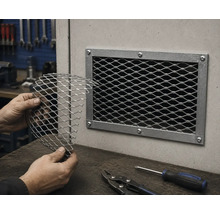Händer som håller ett stycke sträckmetall framför ett monterat ventilationsgaller i en verkstad med plåtsax och skruvmejsel.