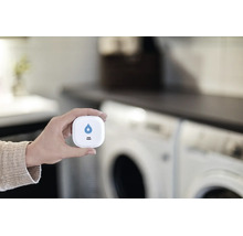 En hand håller en smart vattenläckagesensor framför en tvättmaskin för läckagedetektering i hemmet.
