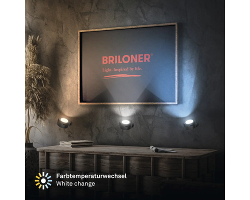 Tre LED väggstrålkastare med justerbar färgtemperatur, monterade under en bild med Briloner logotyp på en texturerad vägg ovanför ett träskåp.