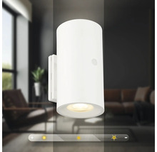 Modern cylindrisk LED vägglampa med dimbar ljusfunktion framför en suddig vardagsrumsbakgrund.