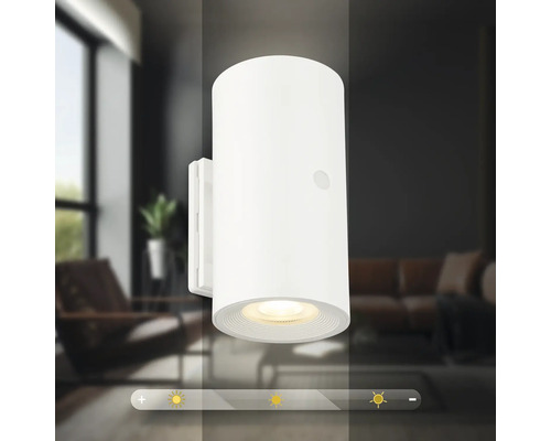 Modern cylindrisk LED vägglampa med dimbar ljusfunktion framför en suddig vardagsrumsbakgrund.
