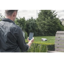 Man med smartphone i trädgården, robotgräsklippare i bakgrunden