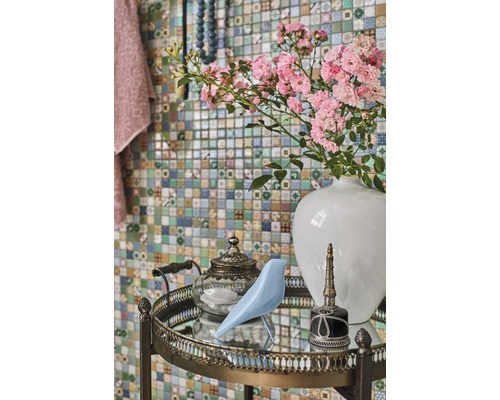 Dekorativt arrangemang med blommor, fågel, tekanna och flaska på ett sidobord framför en mosaikvägg