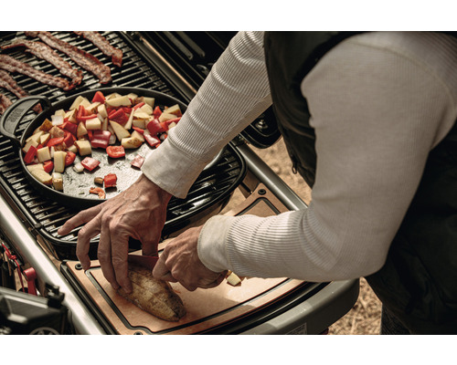 En person förbereder mat på en grill, inklusive bacon, grönsaker i en panna och skivad potatis på en skärbräda.