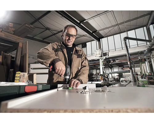 En man monterar beslag på en möbel med en borrmaskin i en verkstad.