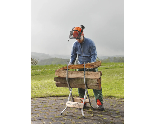 En man med skyddshjälm och visir arbetar med en vedklyv på vilken det ligger vedträn.