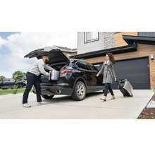 En man packar resväskor i bagageutrymmet på en bil, medan en kvinna drar en bärbar strömgenerator på hjul.