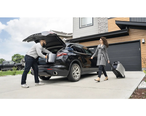 En man packar resväskor i bagageutrymmet på en bil, medan en kvinna drar en bärbar strömgenerator på hjul.