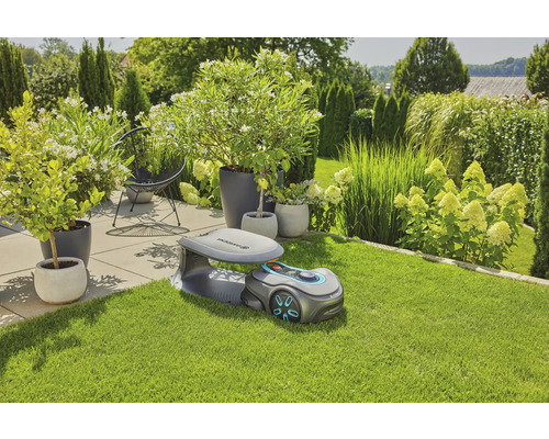 Scen med Gardena robotgräsklippare på gräsmatta, med växter och buskar i bakgrunden.