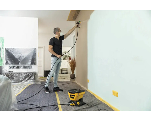 Man målar vägg med färgspraysystem i ett vardagsrum skyddat med täckmaterial.
