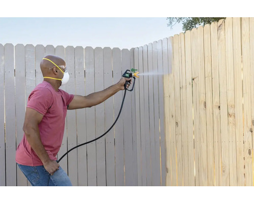 En person målar ett trästaket med en färgspruta, och bär skyddsmask och skyddsglasögon.