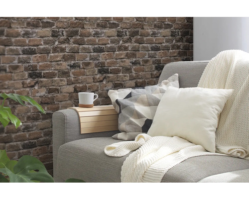 Dekorativt vardagsrum med grå soffa, kuddar, pläd och vägg i tegelstenslook.