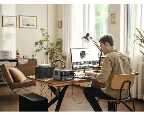 Man arbetar på en dator med en bärbar kraftstation, en router och en nätverkslagringsenhet på ett bord i ett ljust rum.