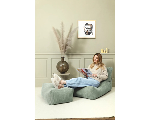 Kvinna läser på en grön manchesterfåtölj med fotpall i ett ljust rum.