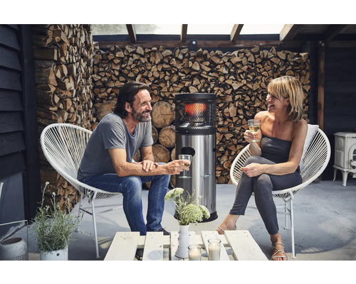 Scen med ett par som sitter på en terrass i vita stolar, med en uteplatsvärmare och ved i bakgrunden.