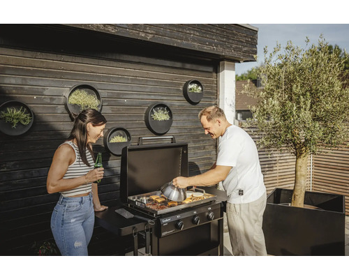 Två personer grillar hamburgare på en Enders gasolgrill utomhus, med runda planteringskärl på väggen.
