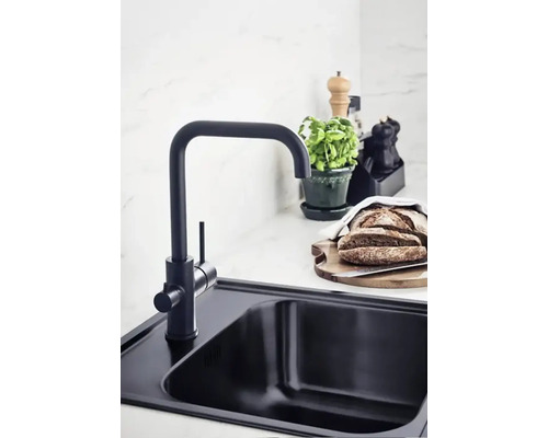 Svart diskho med blandare, skärbräda med bröd, örtkruka och kryddkvarn på en köksbänk.