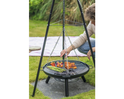 Person grillar på en trebensgrill med ett grillgaller som en kedja är fäst vid. På gallret ligger spett, kött och grön sparris.