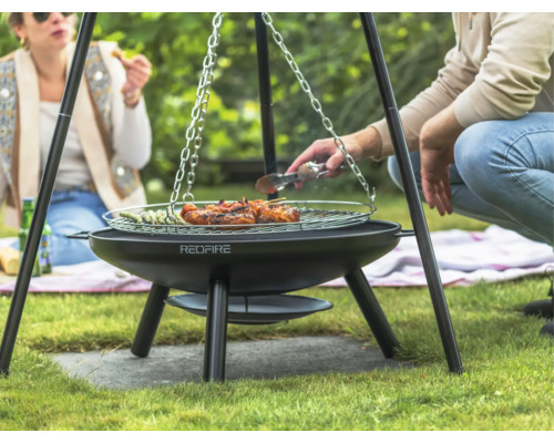 Tre personer grillar kött och grönsaker på en trebensgrill i trädgården.