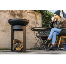 Eldskål i metall med träfack i trädgården och kvinna som dricker en dryck