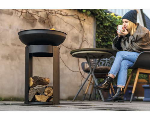 Eldskål i metall med träfack i trädgården och kvinna som dricker en dryck