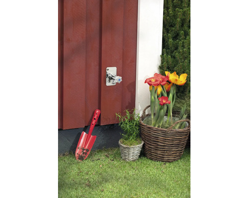 Utomhusbild av en vattenkran, en spade och en blomkorg mot en röd trävägg