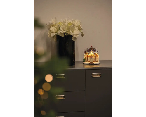 Dekorativ scen med vas med konstgjorda blommor och upplyst pepparkakshus på en byrå.