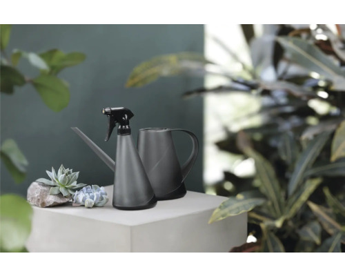 Vattenkanna och växtspruta på piedestal med växter i bakgrunden.