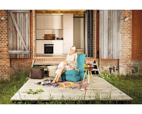 Kvinna sitter på en turkos sittsäck på en träterrass med kuddar och ett sidobord framför en tegelbyggnad med trädörrar.