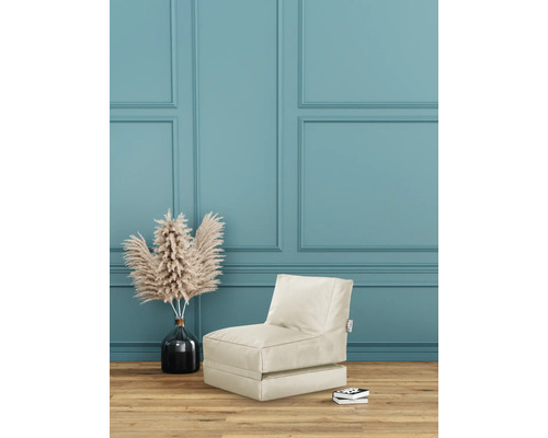 Scen med en beige sittsäcksfåtölj, en vas med torkade blommor och en stapel böcker framför en vägg med paneler