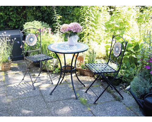 Trädgårdsscen med bord och två stolar med mosaikdetaljer framför frodig grönska